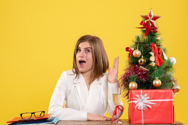 Вид спереди женщина-врач, сидящая перед своим столом на желтом фоне с елкой и подарочными коробками