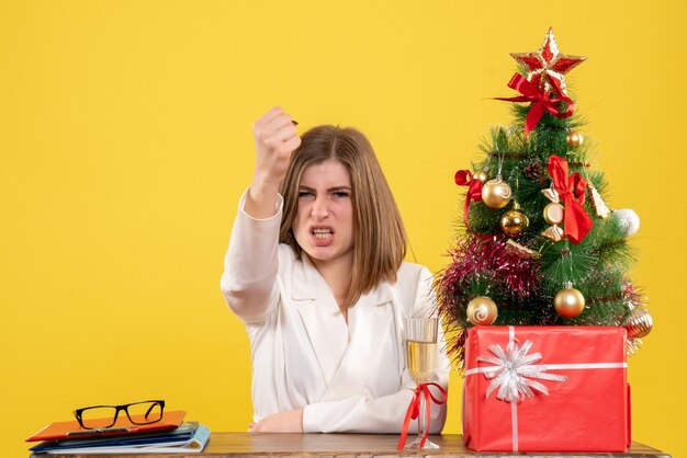 Front view female doctor sitting in front of her table on yellow background with christmas tree and gift boxes