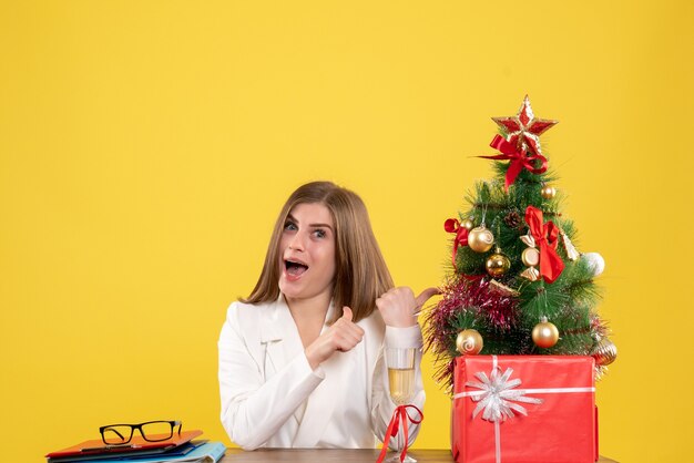 Вид спереди женщина-врач, сидящая перед своим столом на желтом фоне с елкой и подарочными коробками