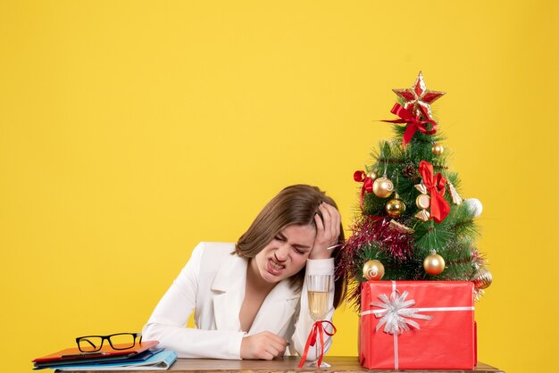 Front view female doctor sitting in front of her table on yellow background with christmas tree and gift boxes