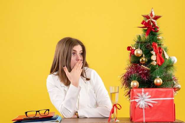 Front view female doctor sitting in front of her table on yellow background with christmas tree and gift boxes