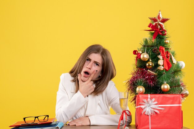 クリスマスツリーとギフトボックスと黄色の背景に彼女のテーブルの前に座っている正面図の女性医師