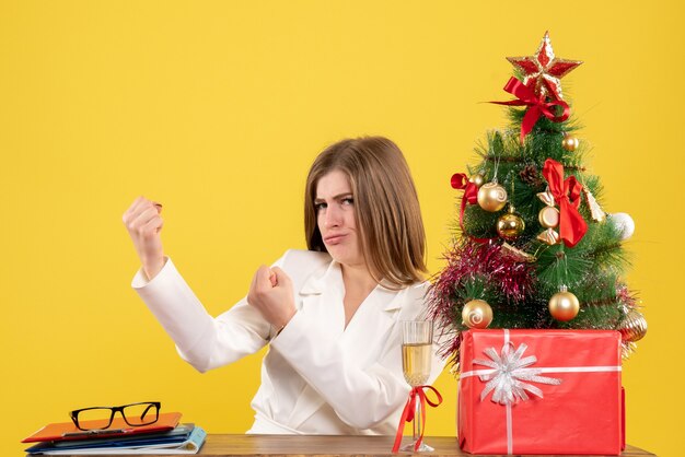 Medico femminile di vista frontale che si siede davanti alla sua tavola sullo scrittorio giallo con i contenitori di regalo e dell'albero di natale