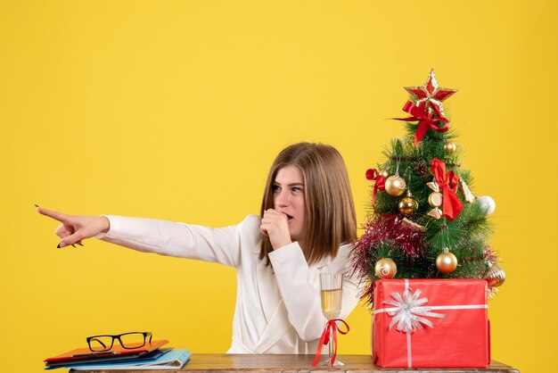 크리스마스 트리와 선물 상자와 노란색 바닥에 그녀의 테이블 앞에 앉아 전면보기 여성 의사