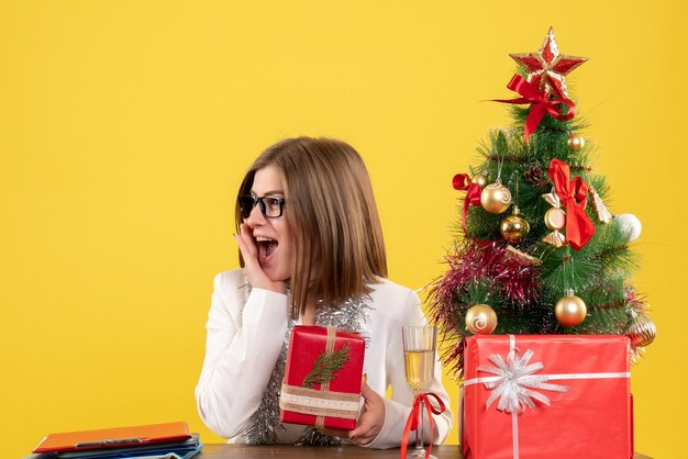 Front view female doctor sitting in front of table with presents and tree on yellow background with christmas tree and gift boxes