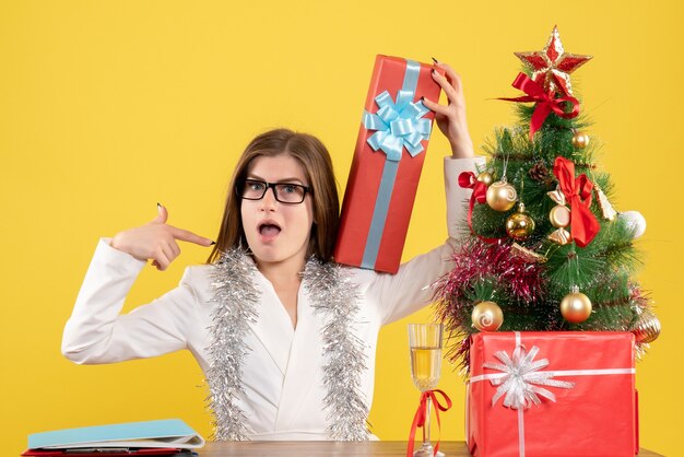 Front view female doctor sitting in front of table with presents and tree on yellow background with christmas tree and gift boxes