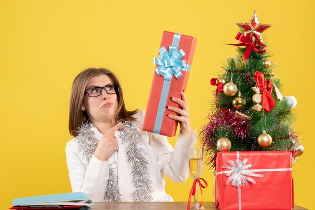 Front view female doctor sitting in front of table with presents and tree on yellow background with christmas tree and gift boxes