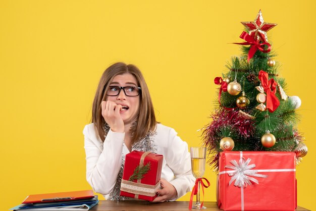 크리스마스 트리 및 선물 상자와 노란색 배경에 선물 및 트리 테이블 앞에 앉아 전면보기 여성 의사