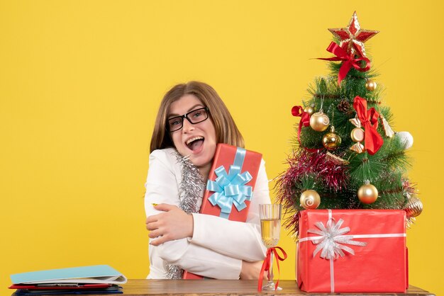Front view female doctor sitting in front of table with presents and tree on yellow background with christmas tree and gift boxes