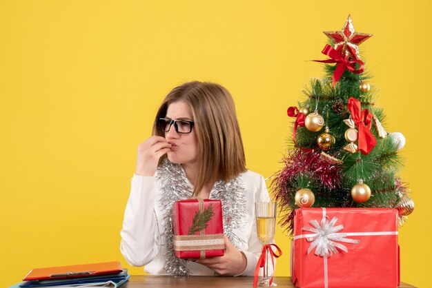 Front view female doctor sitting in front of table with presents and tree on yellow desk