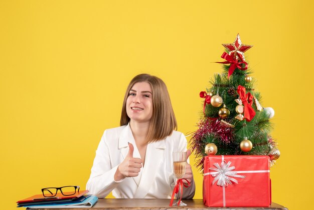 クリスマスプレゼントと黄色の背景に笑顔の木とテーブルの前に座っている正面図の女性医師