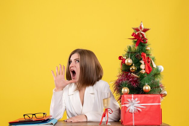 Front view female doctor sitting in front of table on yellow background with christmas tree and gift boxes