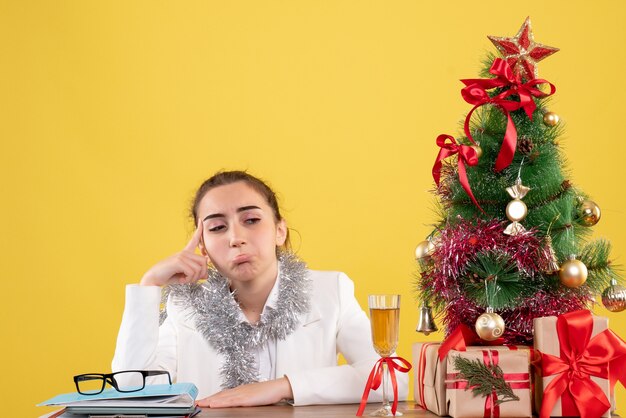 크리스마스 트리와 선물 상자와 노란색 배경에 슬픈 느낌 그녀의 테이블 뒤에 앉아 전면보기 여성 의사