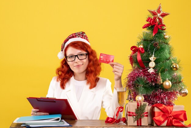 그녀의 테이블 뒤에 앉아 크리스마스 트리와 선물 상자와 노란색 배경에 은행 카드를 들고 전면보기 여성 의사
