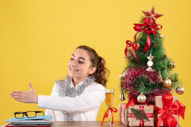 彼女のテーブルの後ろに座って、クリスマスツリーとギフトボックスと黄色の背景に手を振る正面図の女性医師