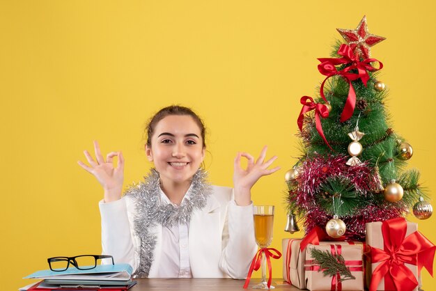 クリスマスツリーとギフトボックスと黄色の背景に笑みを浮かべて彼女のテーブルの後ろに座っている正面図