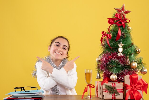 クリスマスツリーとギフトボックスと黄色の背景に笑みを浮かべて彼女のテーブルの後ろに座っている正面図