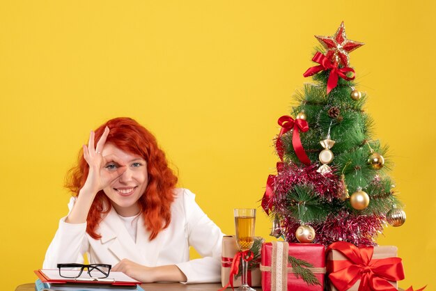 Front view female doctor sitting behind her table with christmas presents and tree smiling on yellow background