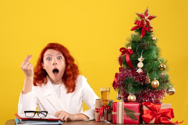 Front view female doctor sitting behind her table with christmas presents and tree on yellow background