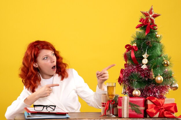 Front view female doctor sitting behind her table with christmas presents and tree on yellow background