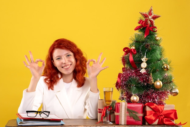 Front view female doctor sitting behind her table with christmas presents and tree on yellow background