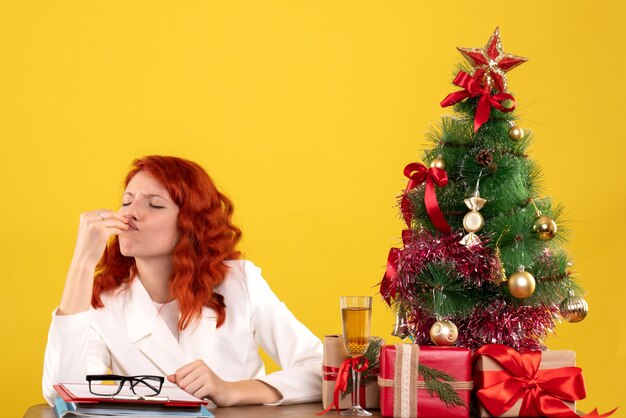 Front view female doctor sitting behind her table with christmas presents and tree on yellow desk