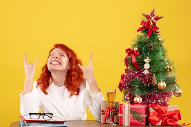 Front view female doctor sitting behind her table with christmas presents and tree on yellow desk