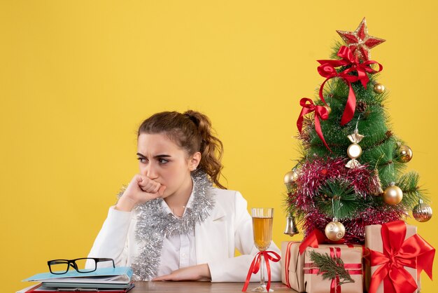 크리스마스 트리와 선물 상자와 노란색 배경에 스트레스 얼굴로 그녀의 테이블 뒤에 앉아 전면보기 여성 의사