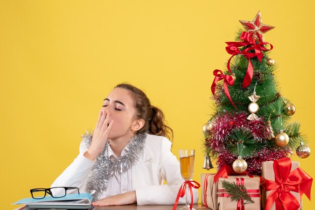 Front view female doctor sitting behind her table and yawning on yellow background with christmas tree and gift boxes