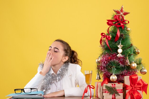 Вид спереди женщина-врач, сидящая за столом и зевая на желтом фоне с рождественской елкой и подарочными коробками