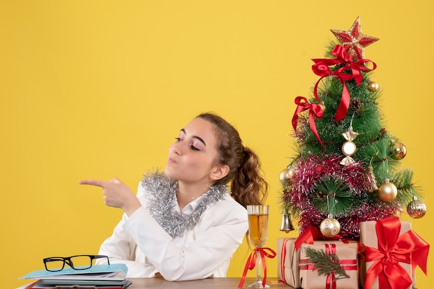 Front view female doctor sitting behind her table on a yellow background with christmas tree and gift boxes