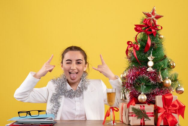 クリスマスツリーとギフトボックスと黄色の背景に彼女のテーブルの後ろに座っている正面図の女性医師