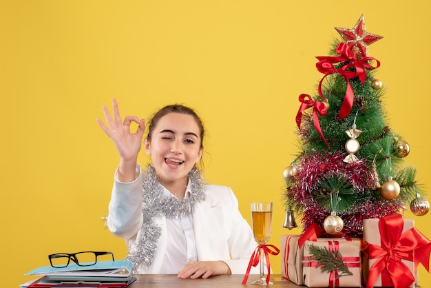 Front view female doctor sitting behind her table on yellow background with christmas tree and gift boxes