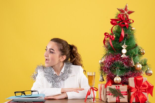 Front view female doctor sitting behind her table on yellow background with christmas tree and gift boxes