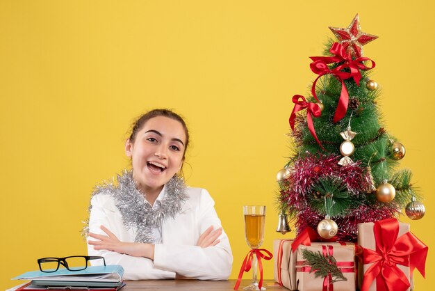 크리스마스 트리와 선물 상자와 노란색 배경에 그녀의 테이블 뒤에 앉아 전면보기 여성 의사