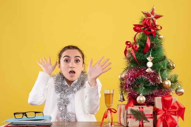 Front view female doctor sitting behind her table on yellow background with christmas tree and gift boxes