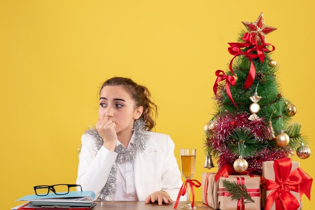 크리스마스 트리와 선물 상자와 노란색 배경에 그녀의 테이블 뒤에 앉아 전면보기 여성 의사