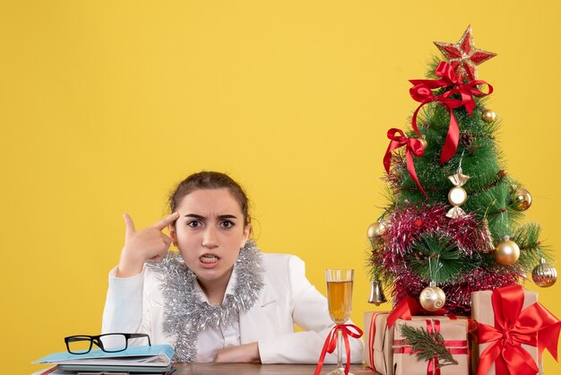 Front view female doctor sitting behind her table on yellow background with christmas tree and gift boxes