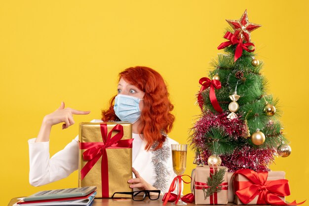 Front view female doctor sitting in mask with xmas presents and tree on yellow background