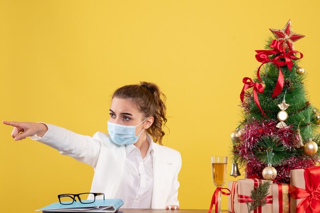 Front view female doctor sitting in protective mask pointing on yellow background