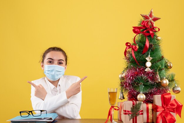 クリスマスツリーとギフトボックスと黄色の背景に笑みを浮かべて保護マスクに座っている正面図女性医師