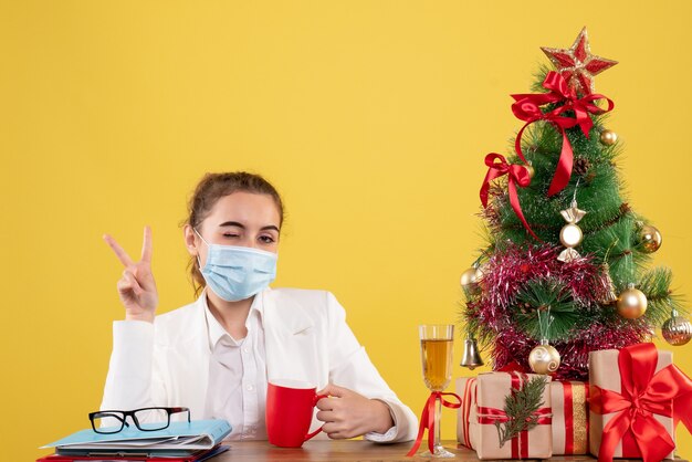 크리스마스 트리와 선물 상자와 노란색 배경에 보호 마스크에 앉아 전면보기 여성 의사