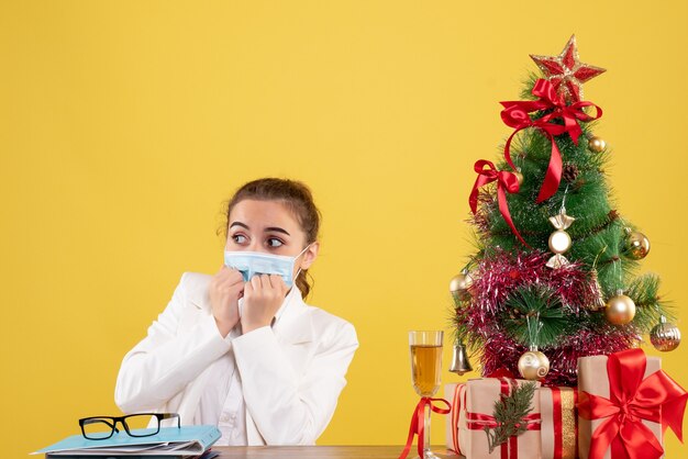 Front view female doctor sitting in protective mask on the yellow background with christmas tree and gift boxes