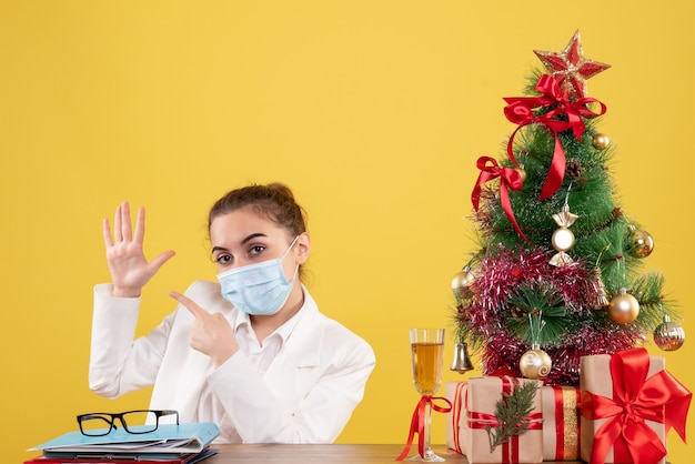 크리스마스 트리와 선물 상자와 노란색 배경에 보호 마스크에 앉아 전면보기 여성 의사