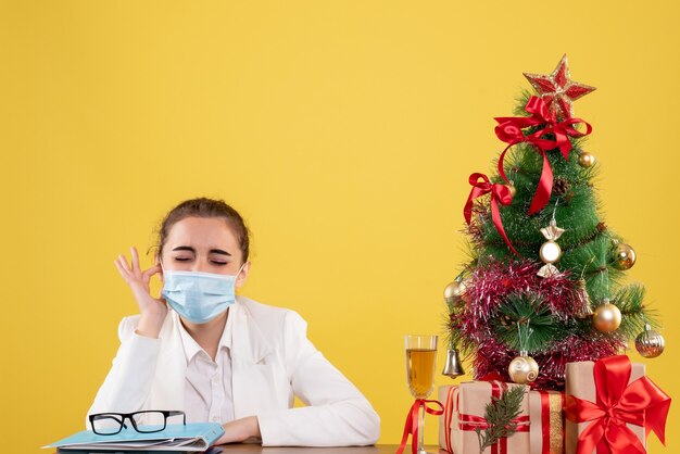 Front view female doctor sitting in protective mask on yellow background with christmas tree and gift boxes