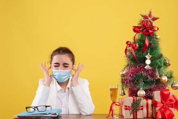 クリスマスツリーとギフトボックスと黄色の背景の保護マスクに座っている正面図女性医師