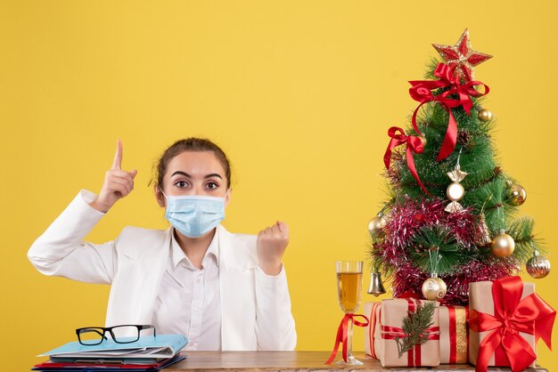 Front view female doctor sitting in protective mask on yellow background with christmas tree and gift boxes