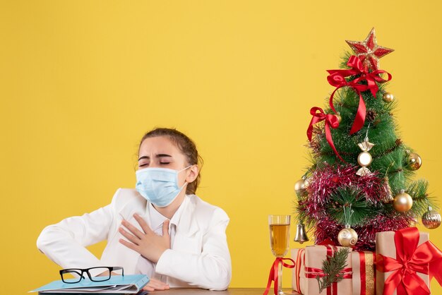 Front view female doctor sitting in protective mask on yellow background with christmas tree and gift boxes