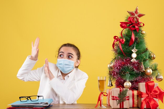 Front view female doctor sitting in sterile mask on the yellow background with christmas tree and gift boxes