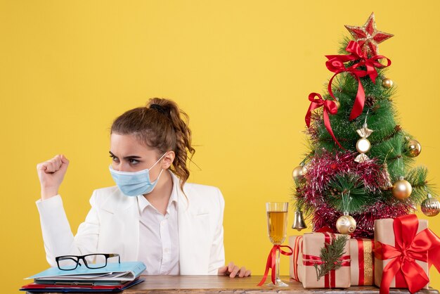 クリスマスツリーとギフトボックスと黄色の背景に滅菌マスクに座っている正面図の女性医師